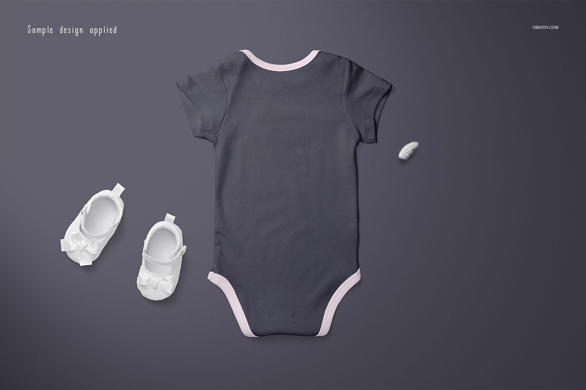Baby onesie mockup in dark gray with light pink trim, displayed flat on a gray surface alongside white baby shoes.