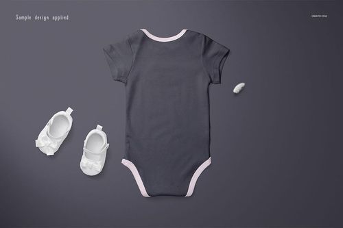 Baby onesie mockup in dark gray with light pink trim, displayed flat on a gray surface alongside white baby shoes.