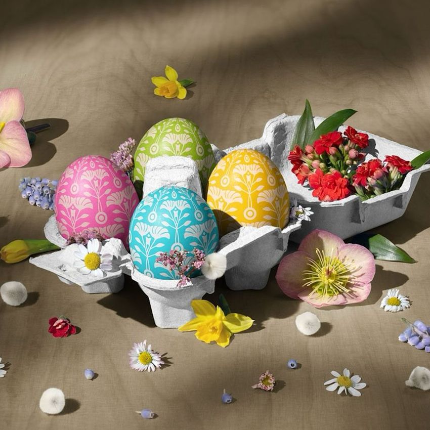 Egg carton mockup with patterned pastel eggs and spring flowers arranged on a brown surface in soft natural light.