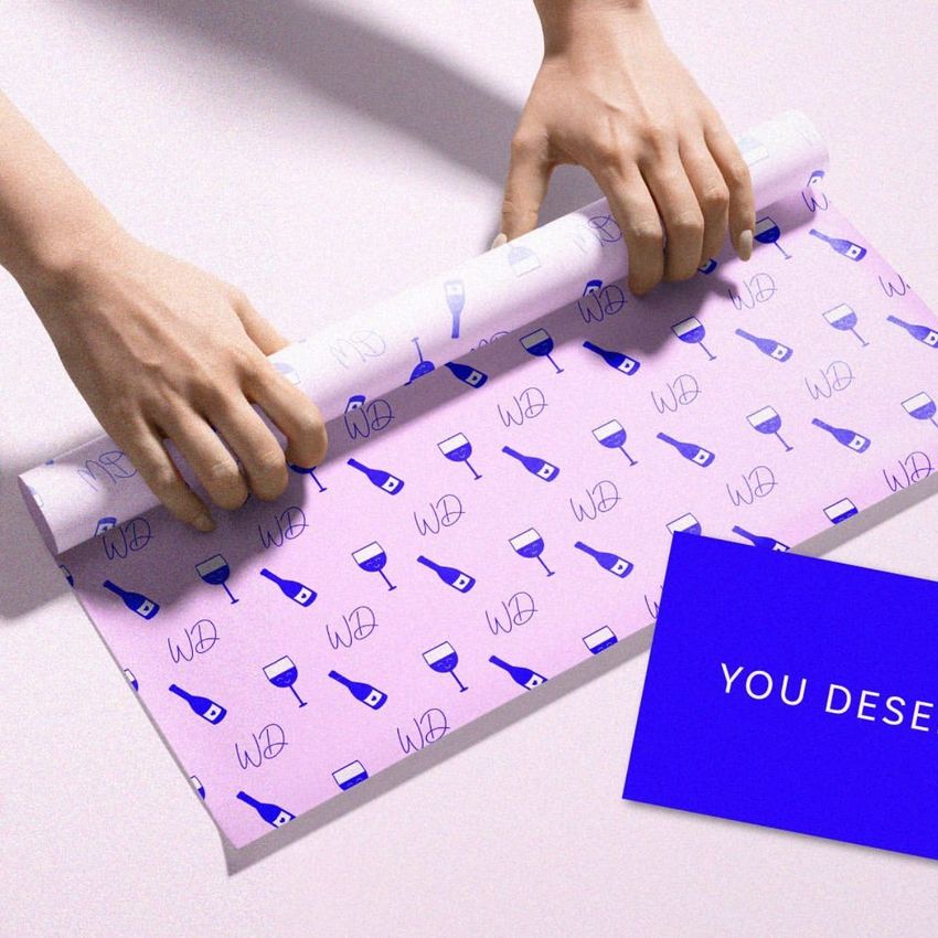 Hands rolling a sheet of food safe paper mockup with a pink and blue wine-themed pattern, next to a blue card with text.