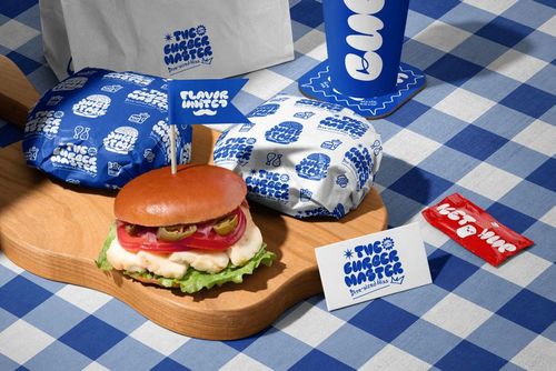 PSD mockup featuring a vegan burger, packaging elements, and a blue cup on a wooden board and checkered cloth.