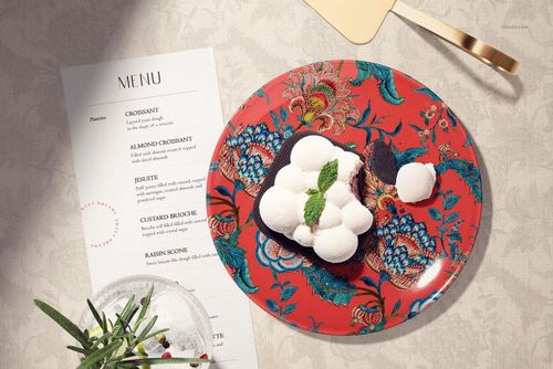 Overhead shot of a patterned ceramic plate mockup featuring a dessert, menu, and utensils on a neutral surface.