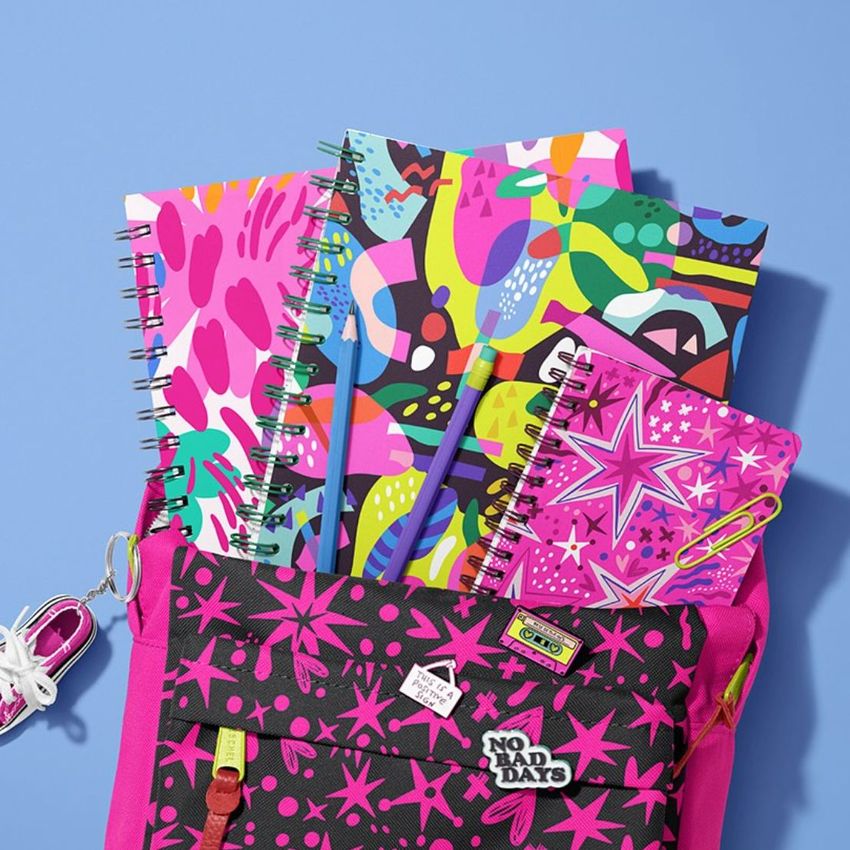 School backpack mockup with spiral notebooks featuring colorful abstract and star patterns, arranged on a light blue surface.