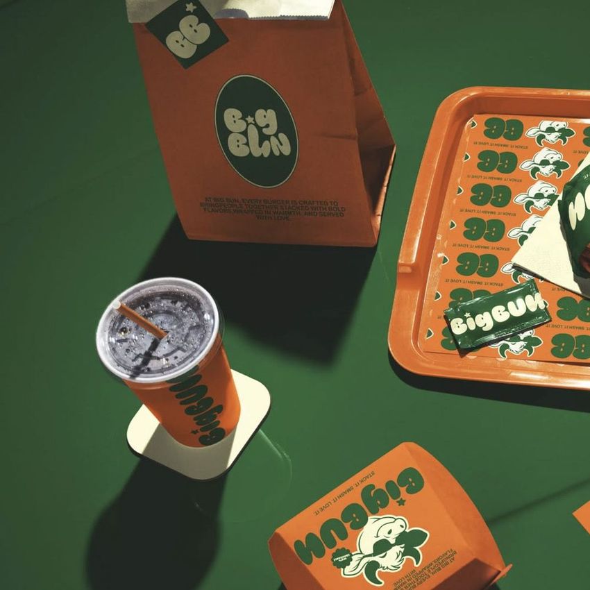 Fast food burger branding mockup featuring orange packaging, drink cup, tray, and paper bag on a green car hood surface.
