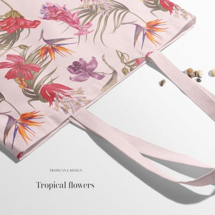 Tote bag mockup featuring a pastel pink fabric with a colorful tropical flower pattern, shown on a light surface with small pebbles nearby.