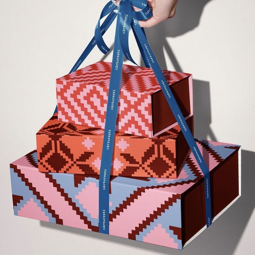 Stacked packaging mockup featuring hand-held gift boxes with bold pink, red, and blue patterns and a blue ribbon.