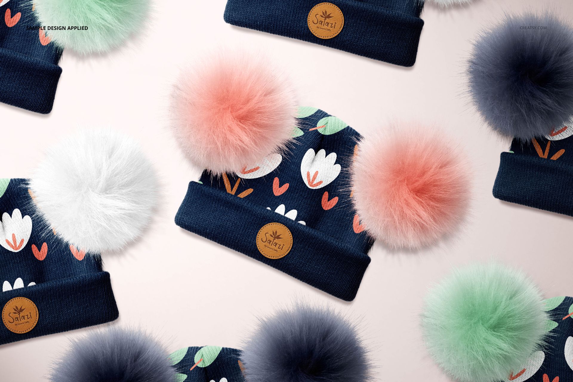 Beanie mockup with large fur pompons in pastel colors, navy blue fabric, floral pattern, top view on light background.