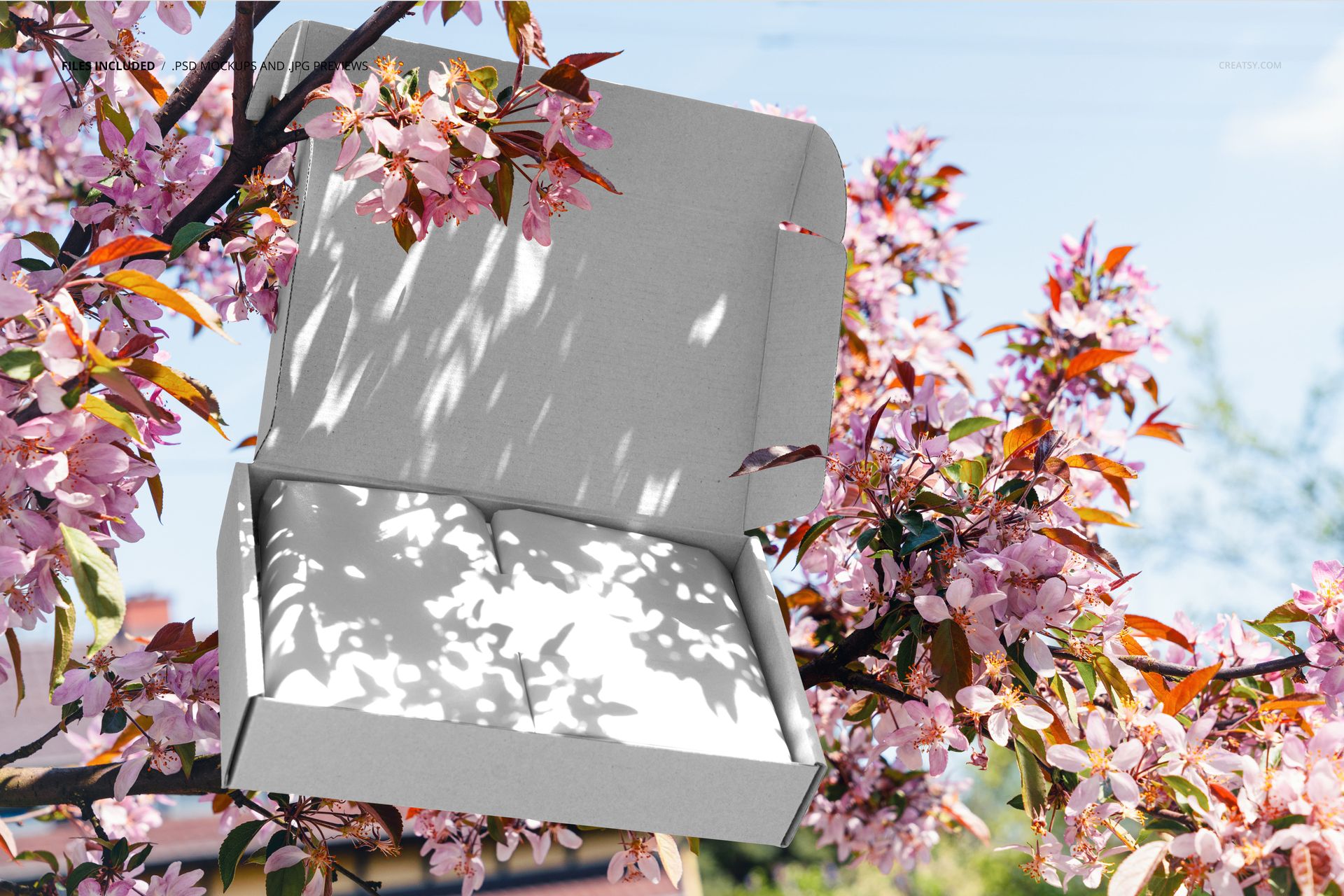 Rectangular mailing box mockup with open lid, placed among blooming pink tree branches in natural daylight.