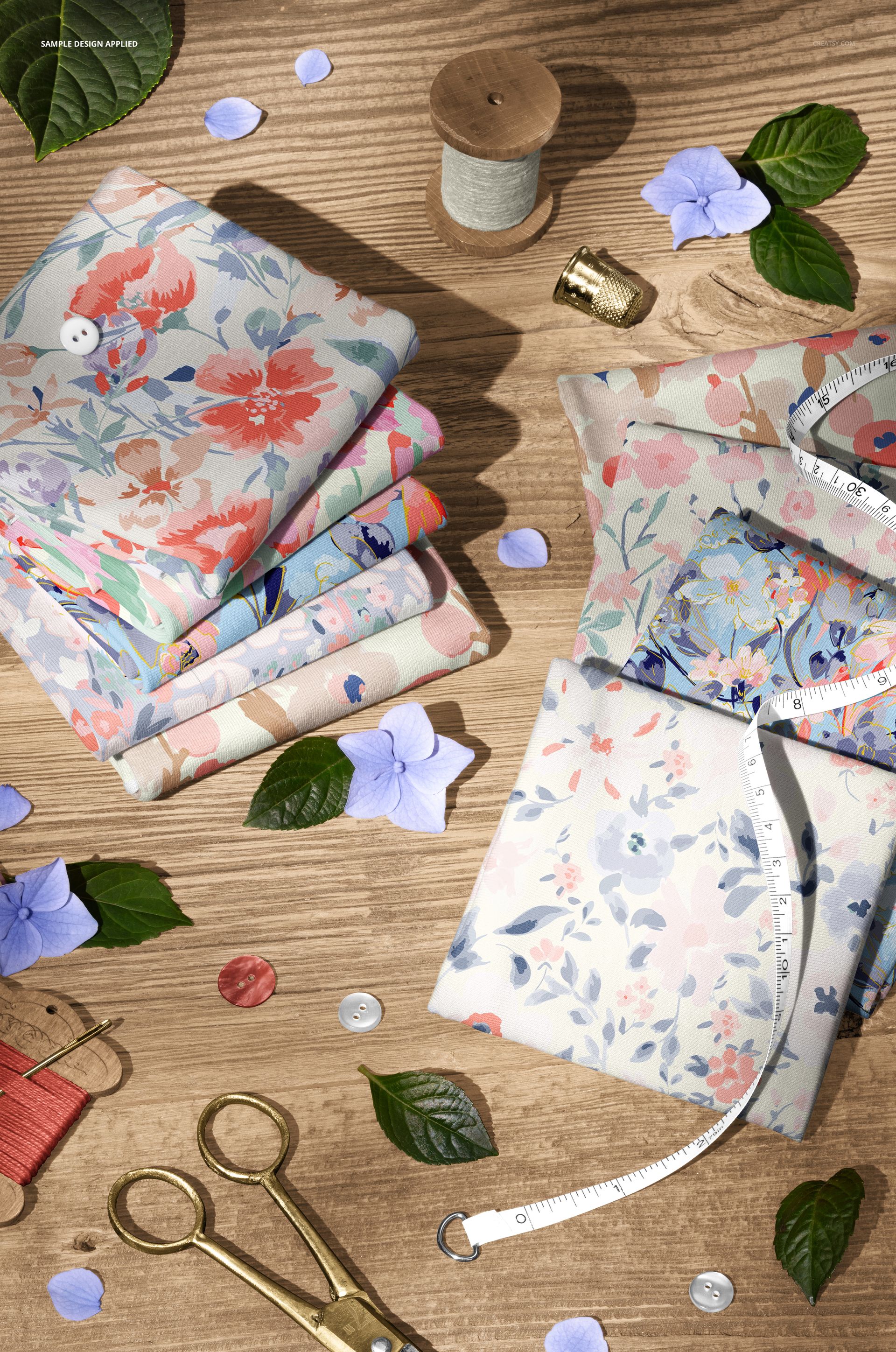 Jersey fabric mockup featuring folded textiles with soft floral designs, displayed on wood with leaves and sewing tools.