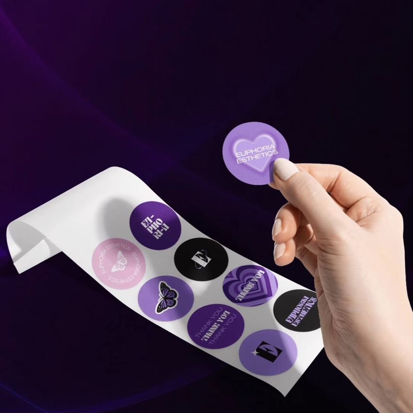 Hand holding a round purple sticker above a sheet of assorted circular stickers in purple, black, and white on a dark background.