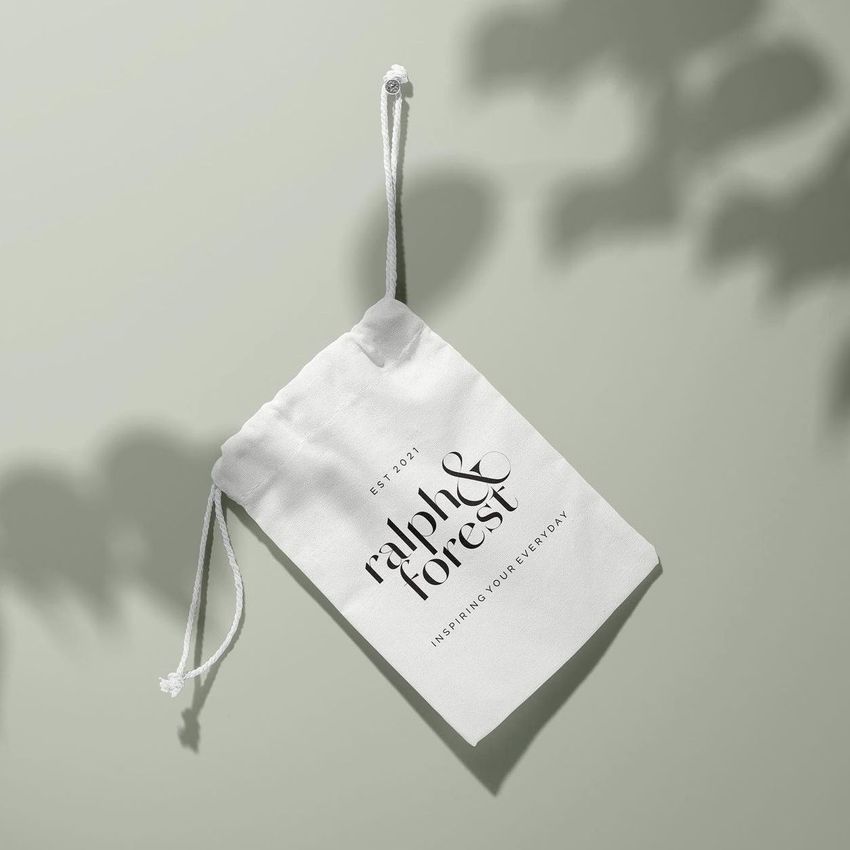 Hanging drawstring pouch mockup featuring a white fabric bag with black text, set on a light green surface with soft shadows.