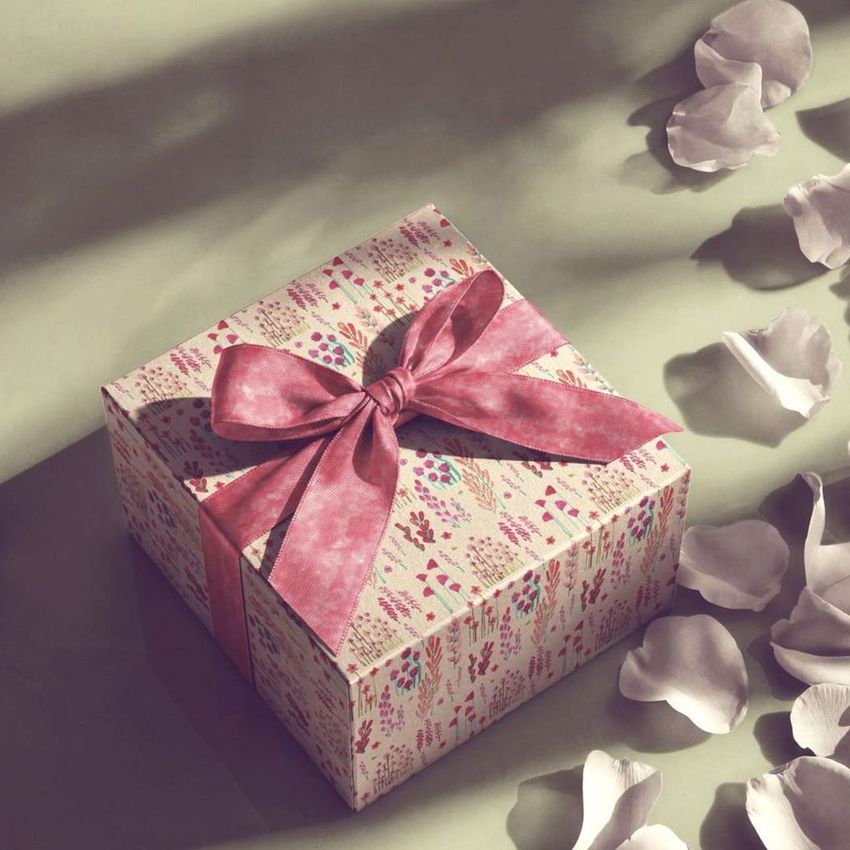 Square gift box mockup with a pink patterned ribbon, surrounded by scattered white petals on a soft green surface.