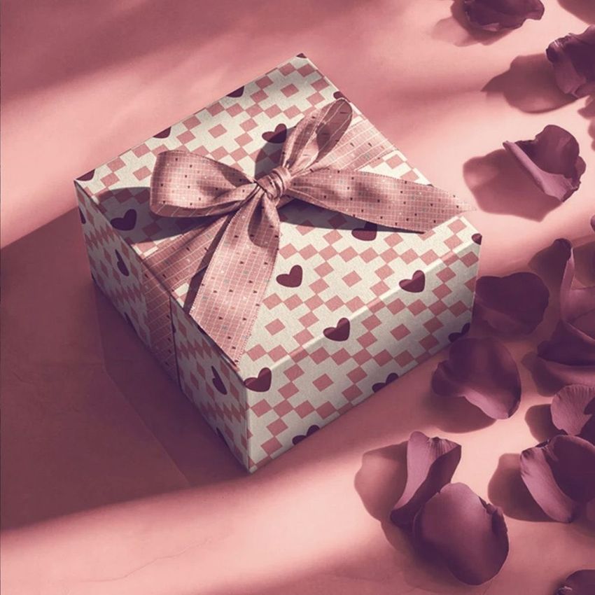 Square gift box mockup with a patterned lid, pink ribbon bow, and scattered rose petals on a pink surface in soft lighting.