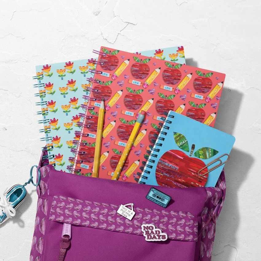 School backpack mockup with spiral notebooks featuring colorful apple and pencil patterns, viewed from above on a light surface.