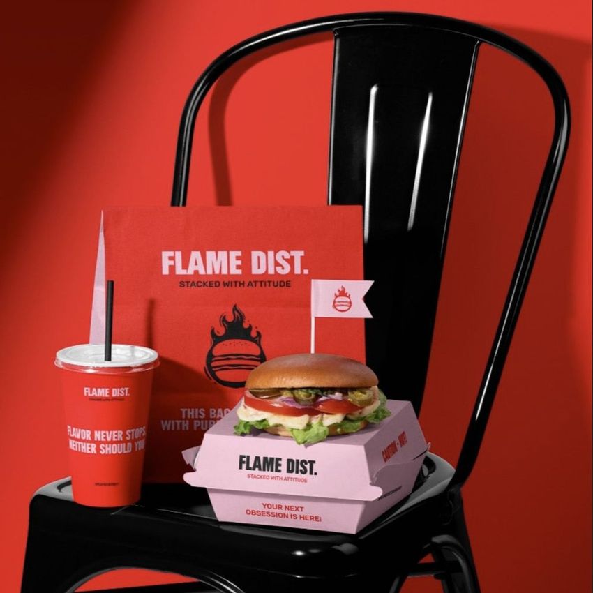 Packaging mockup featuring a vegan burger in a branded box, cup, and paper bag, arranged on a black chair against a red background.