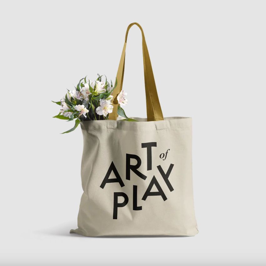 Tote bag mockup featuring a beige fabric bag with bold black text, yellow handles, and white flowers peeking out from the top.