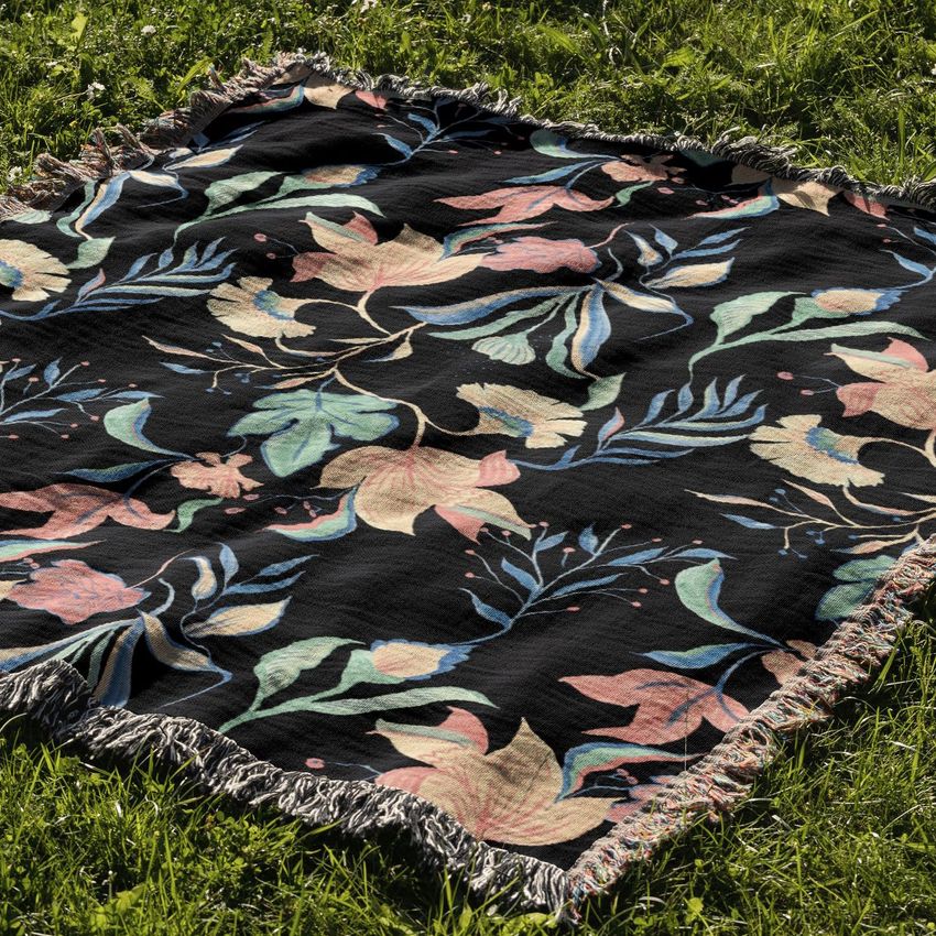 Jacquard fringed woven throw blanket mockup featuring a floral pattern with pastel leaves and flowers on a black background, displayed on grass.