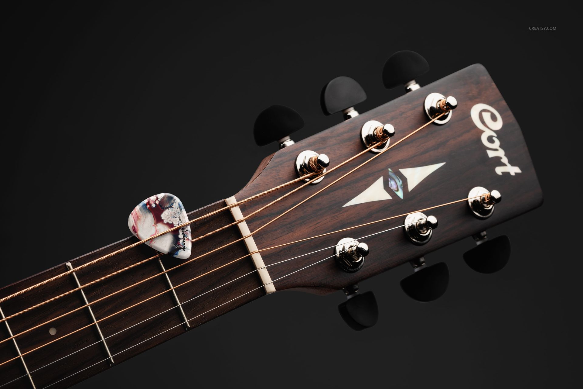 Guitar pick with marbled pattern resting between strings on the headstock of a wooden acoustic guitar, front view.