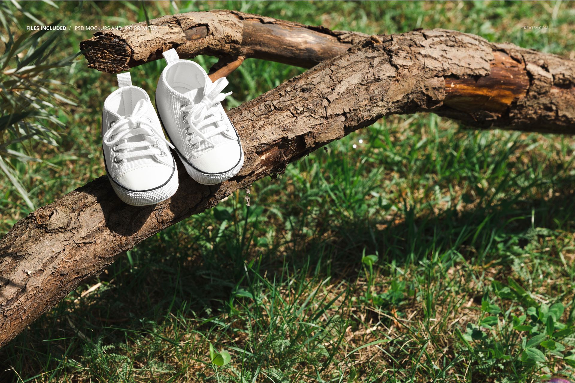 White baby sneakers mockup displayed on a tree branch outdoors, surrounded by green grass and natural sunlight.