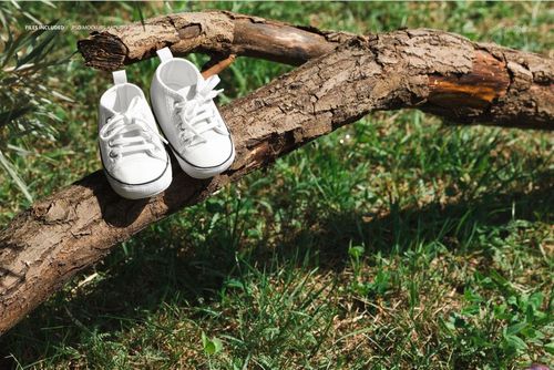 White baby sneakers mockup displayed on a tree branch outdoors, surrounded by green grass and natural sunlight.