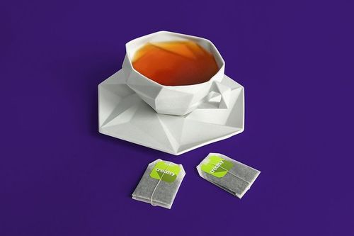 Geometric white tea cup filled with tea on matching saucer, two labeled tea bags on a solid purple background.