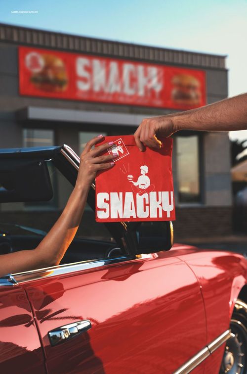 Red fast food paper bag with bold white text being handed to a person in a red convertible at a drive-thru window.