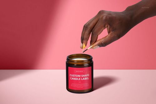 Amber jar candle mockup featuring a custom label, wooden wick, and a hand holding a match on a pink backdrop.