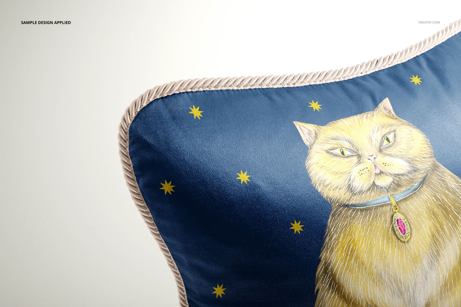Close-up of a silk cushion mockup with braided trim, showing a cat drawing and star pattern on blue fabric.