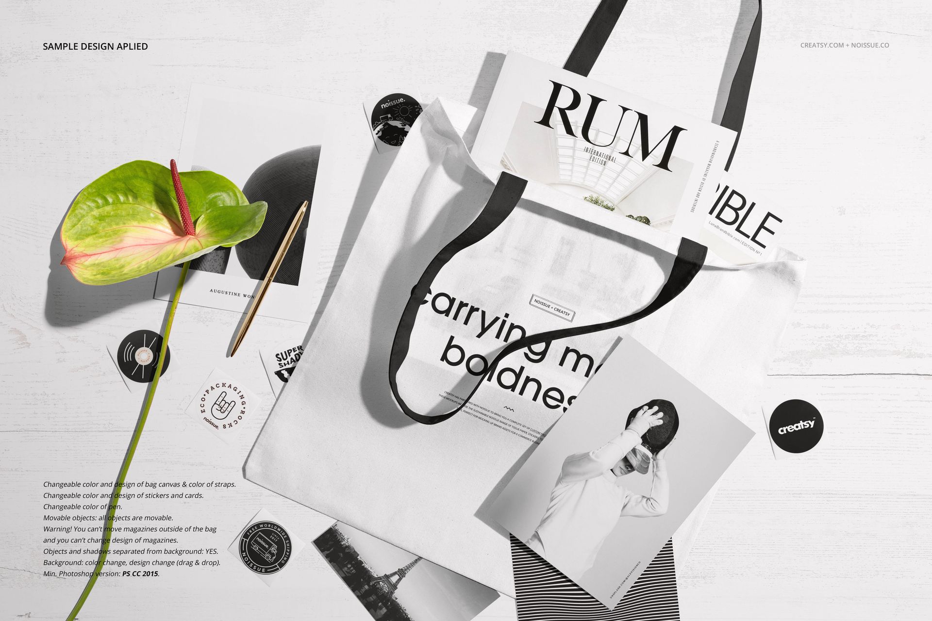 White tote bag mockup displayed on a flat surface with magazines, a pen, a green anthurium flower, and black stickers.