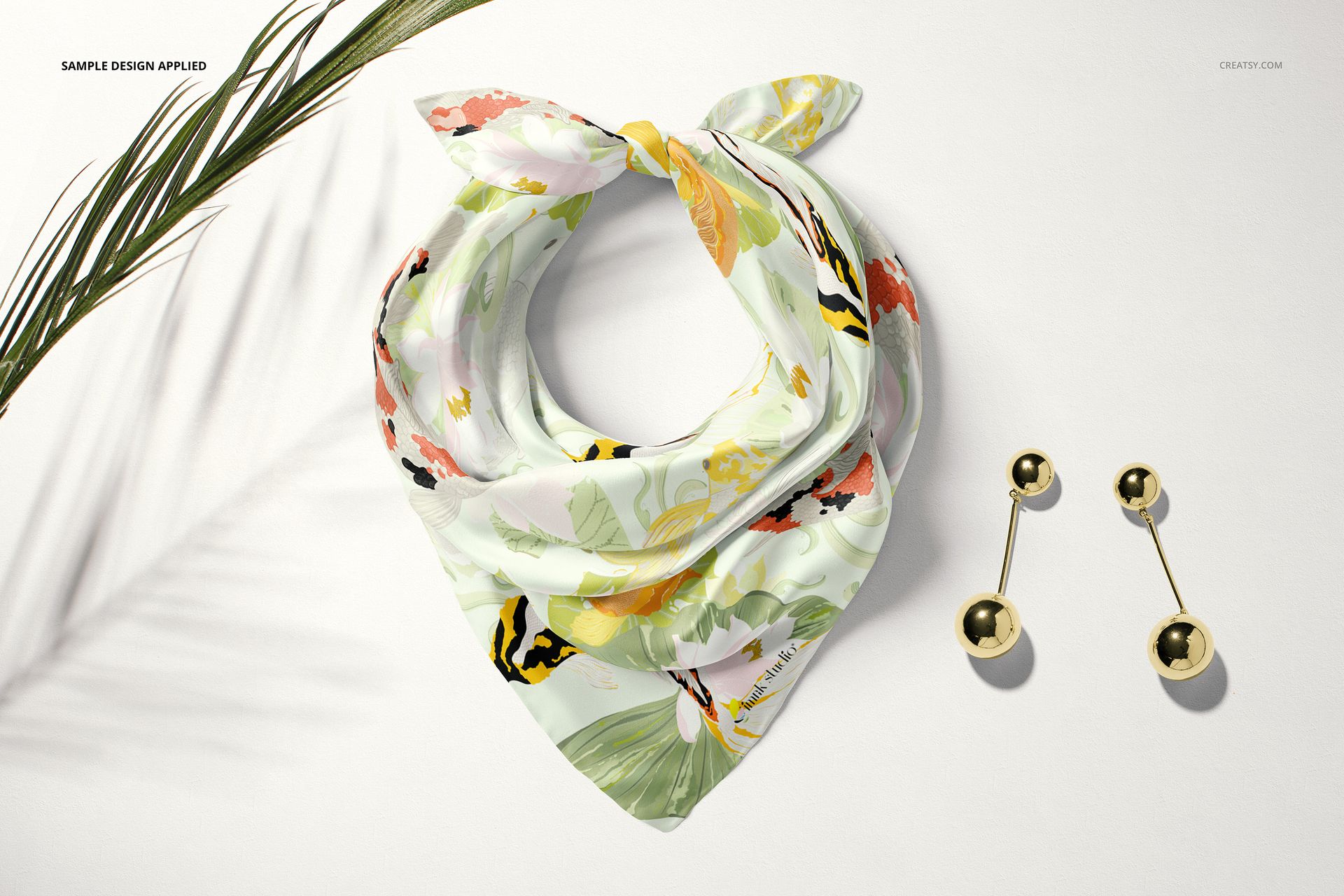 Silk scarf mockup in a top view, featuring soft green and orange floral prints, next to gold ball earrings.