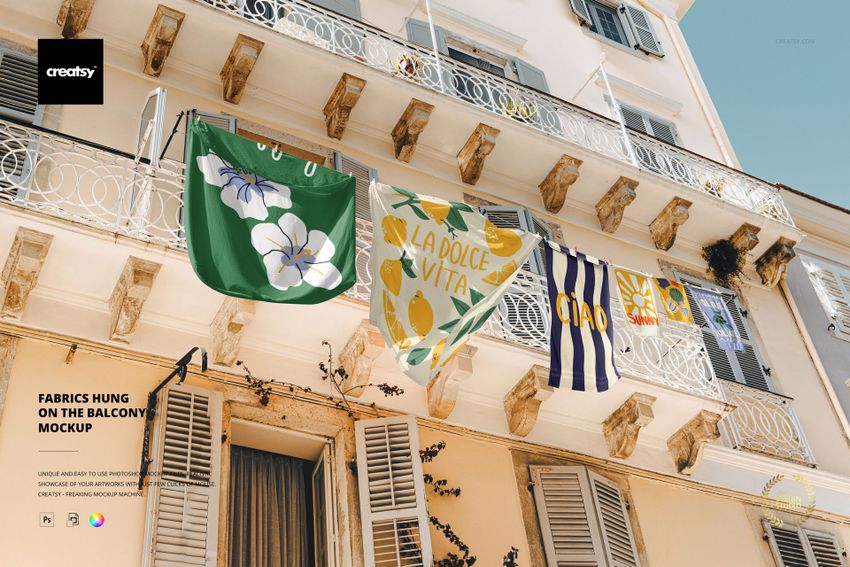Colorful fabrics with floral and striped patterns are hung over a white balcony railing on a sunny building facade.