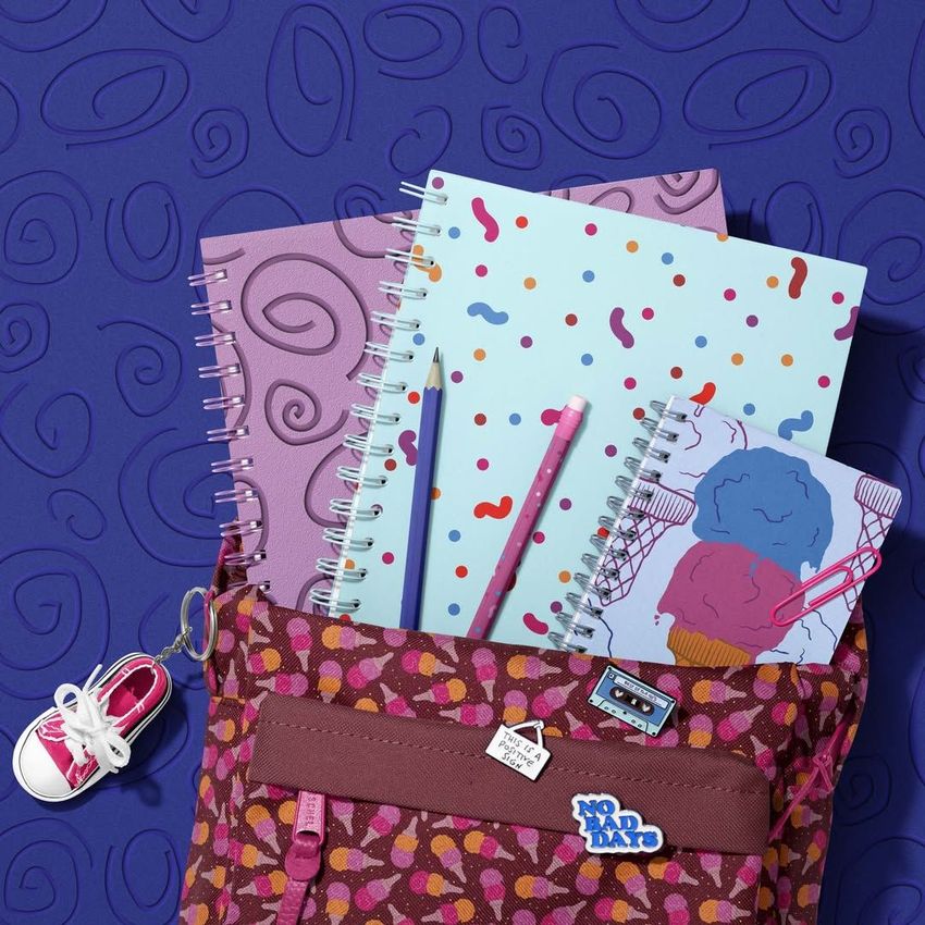 School backpack mockup with spiral notebooks, pencils, and a small sneaker keychain on a purple patterned surface.