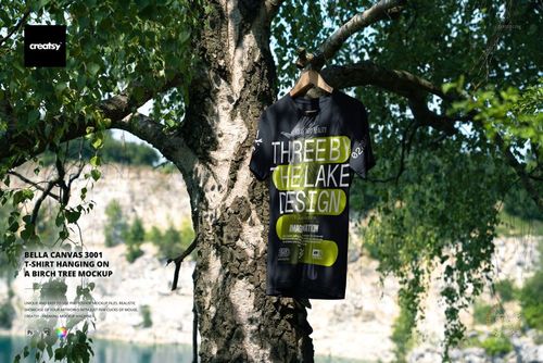 Black adult t-shirt hanging on a birch tree outdoors, surrounded by green leaves, front view mockup scene.