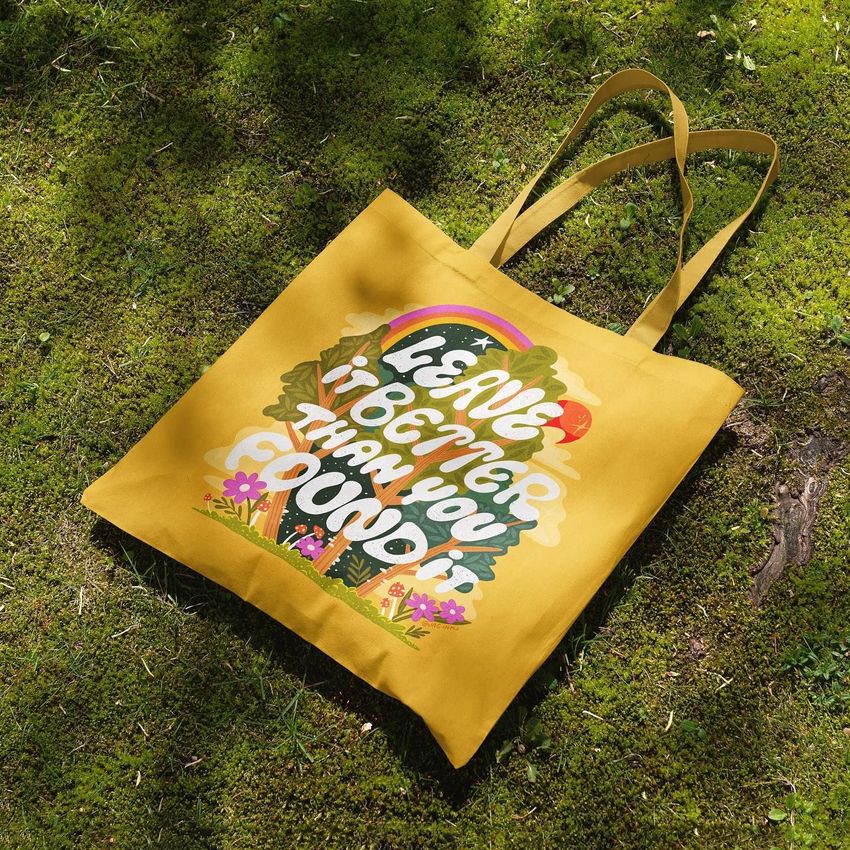 Tote bag mockup featuring a yellow fabric bag with colorful text and nature-themed illustrations, placed on green moss outdoors.