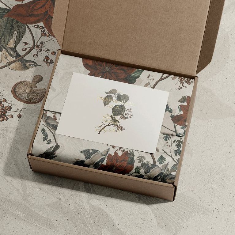 Stationery mockup showing an open kraft box with a floral patterned card and matching wrapping paper inside on a textured surface.