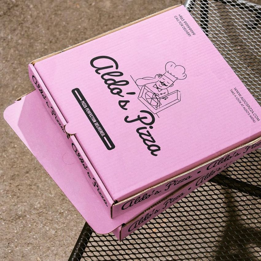 Pink pizza box mockup with illustrated chef and bold text on the lid, placed on an outdoor metal table with a concrete background.