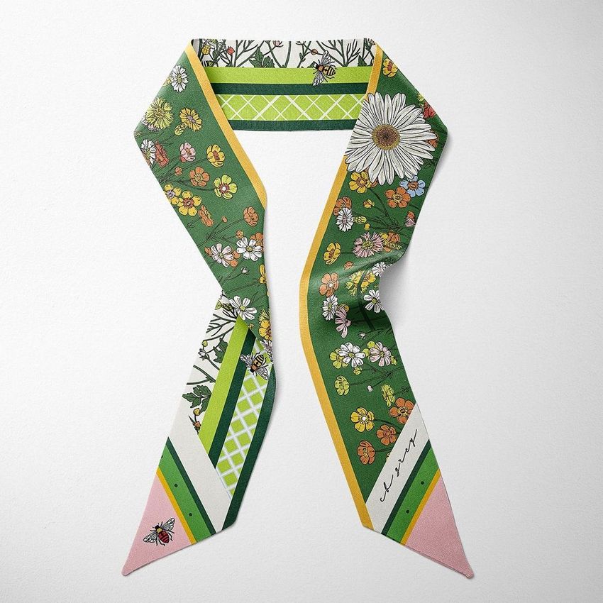 Top view of a twilly scarf mockup featuring a green floral pattern with daisies, bees, and geometric accents on a white surface.