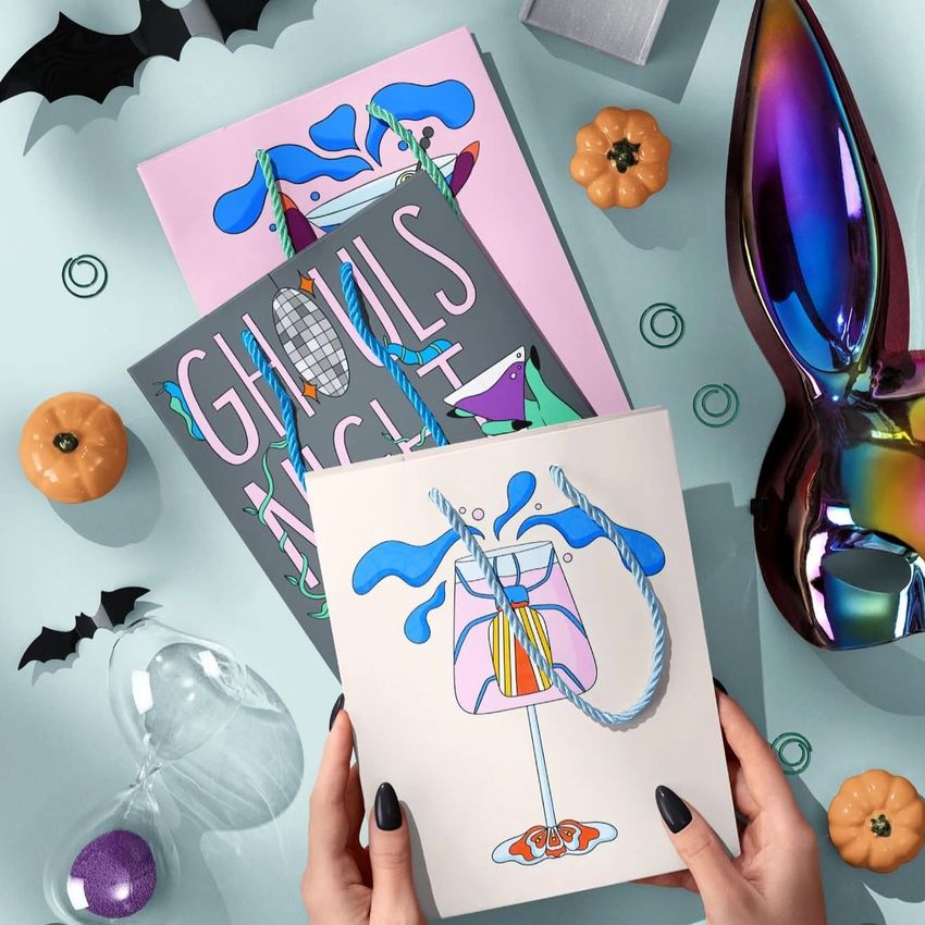 Halloween paper gift bag mockup with illustrated drink and blue handles, surrounded by pumpkins, bats, and festive props on a light blue surface.