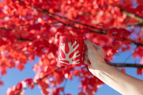Hand holding a red mug with a white abstract pattern outdoors, with vibrant red autumn leaves and blue sky in background.