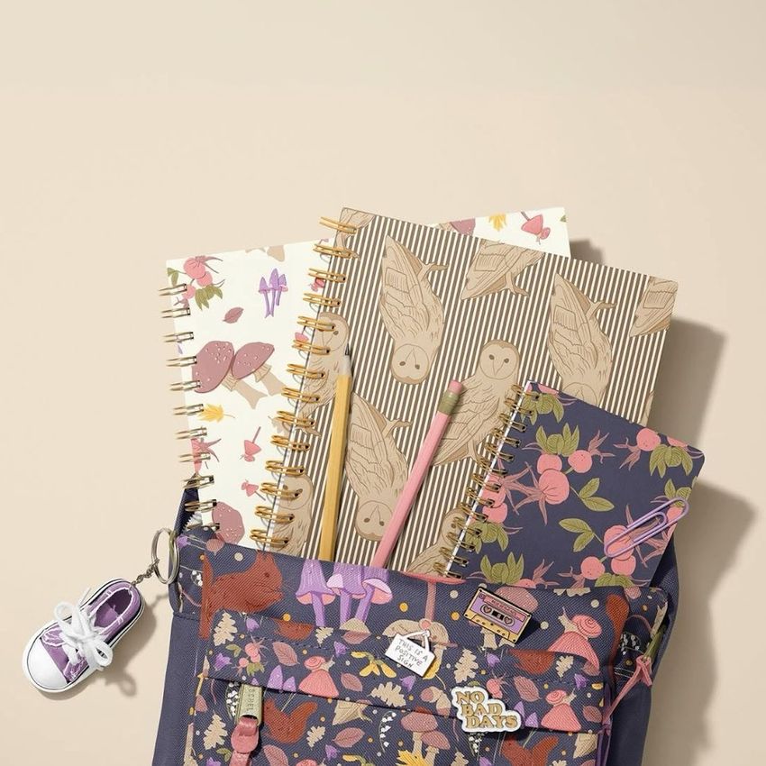 School backpack mockup with woodland animal pattern, spiral notebooks with playful designs, and colored pencils on a beige background.