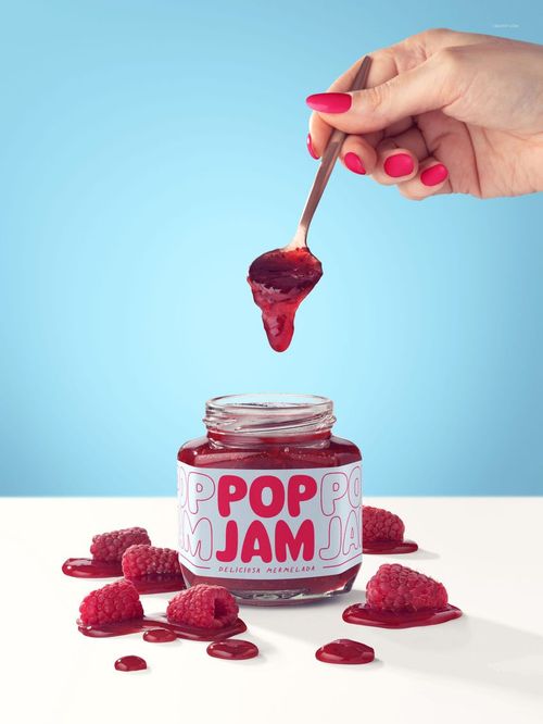 Raspberry jam jar mockup with spoon held above, jam dripping, raspberries and spilled jam on white surface.