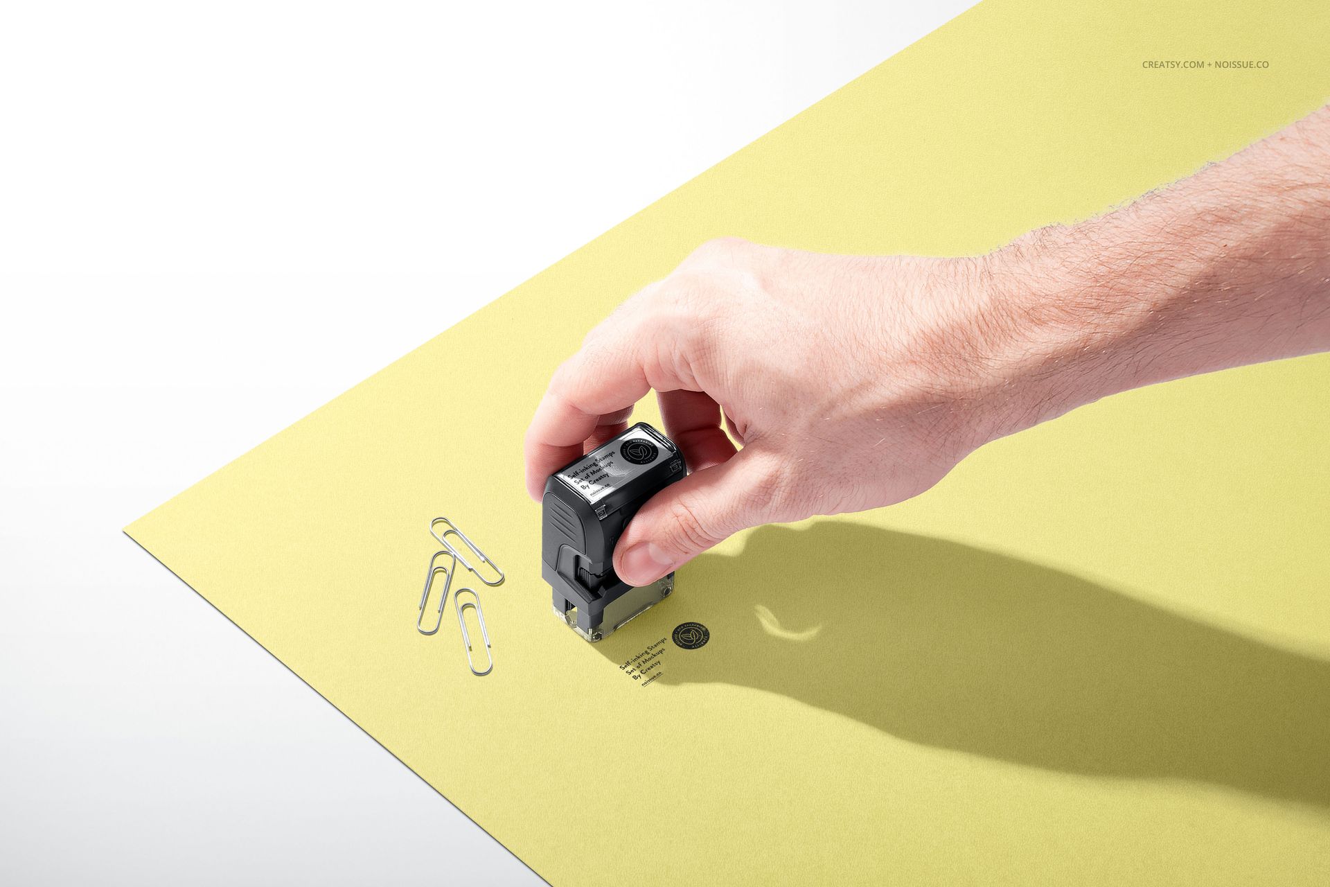 Hand using a self-inking stamp on a yellow sheet of paper with paper clips, shown in a stationery mockup scene.