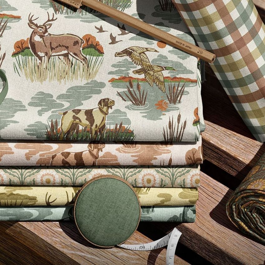 Fabric rolls and stacked fabric pieces with nature-themed patterns on a wooden garden bench, featuring earthy colors and a measuring tape.
