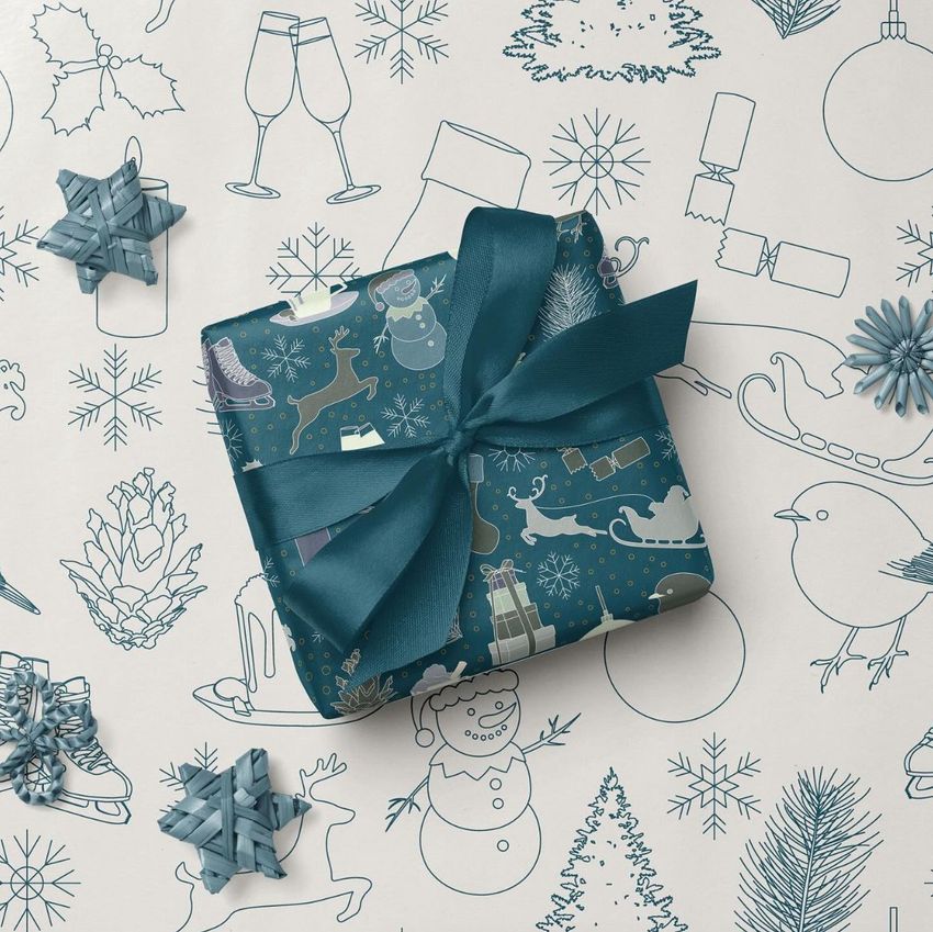 Square gift box wrapped in blue Christmas-themed paper with a matching ribbon, placed on festive patterned wrapping paper.
