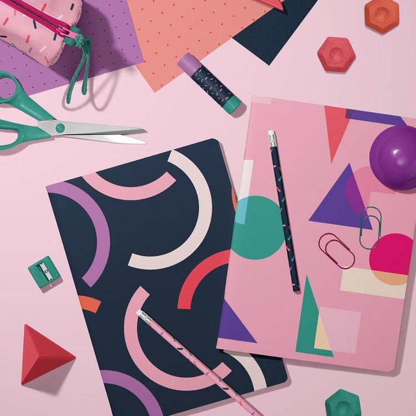 Stationery mockup featuring two A4 notebooks with bold geometric patterns, pencils, paper clips, and colorful desk accessories on a pink surface.