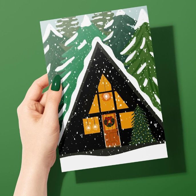 Woman's hand with green nails holding a greeting card mockup featuring a snowy cabin illustration with pine trees on a green background.