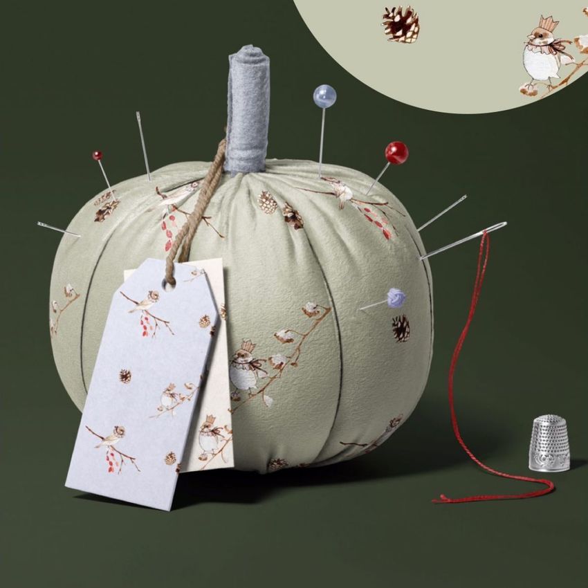 Fabric pumpkin mockup with a light sage green textile featuring bird and pinecone illustrations, used as a pincushion with sewing pins and tags, set against a dark green background.