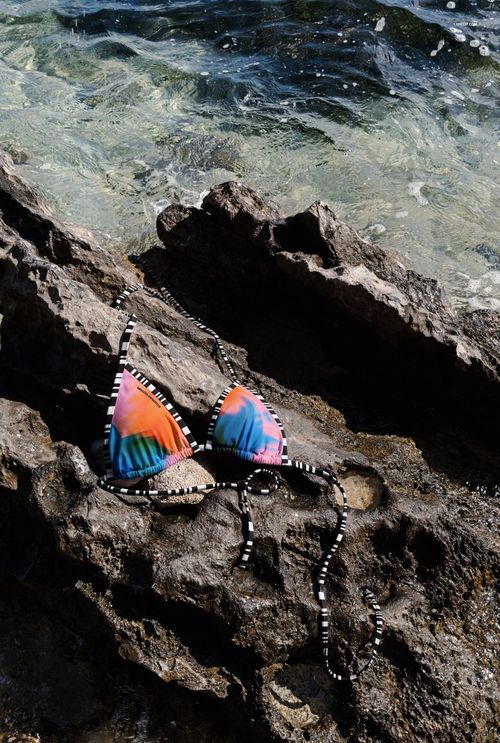 Bikini mockup featuring a vibrant top and bottom with black and white straps, set on rugged rocks by the sea.