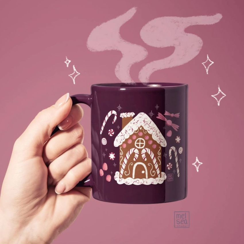 Hand holding a dark purple mug mockup with a gingerbread house and candy illustration, set against a mauve background with drawn steam.