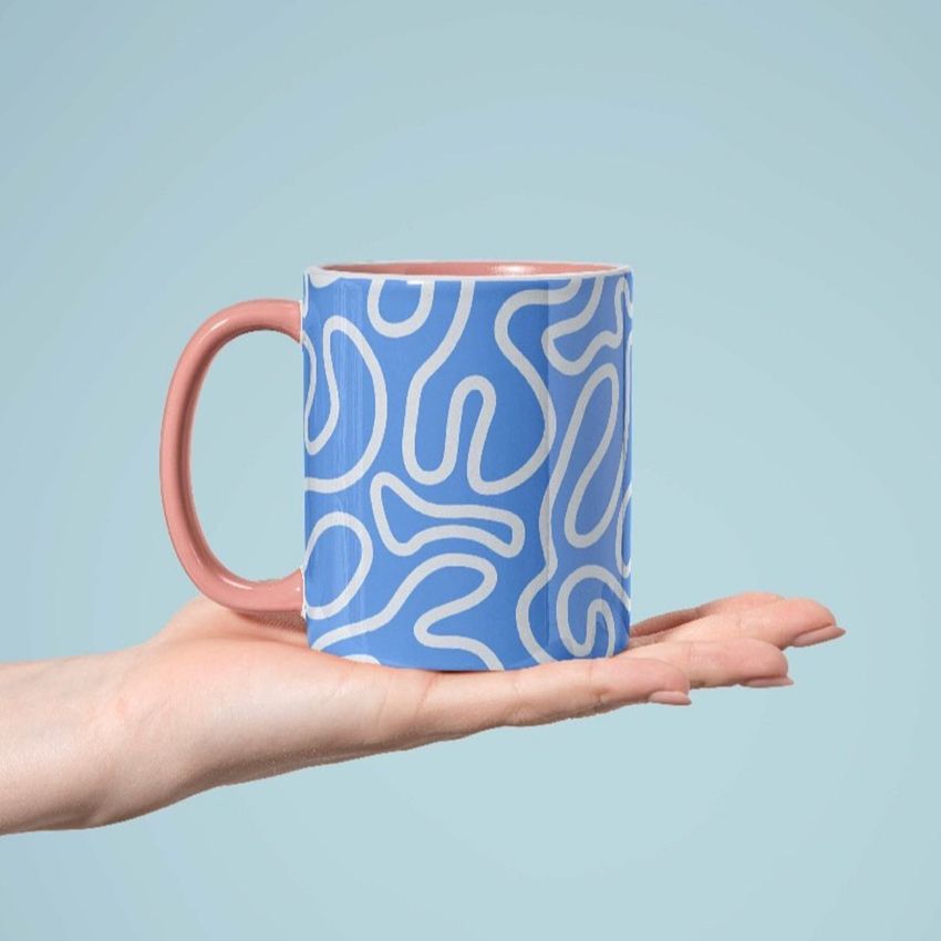 Mug mockup featuring a blue coffee mug with abstract white wave patterns, held on an outstretched hand against a light blue background.